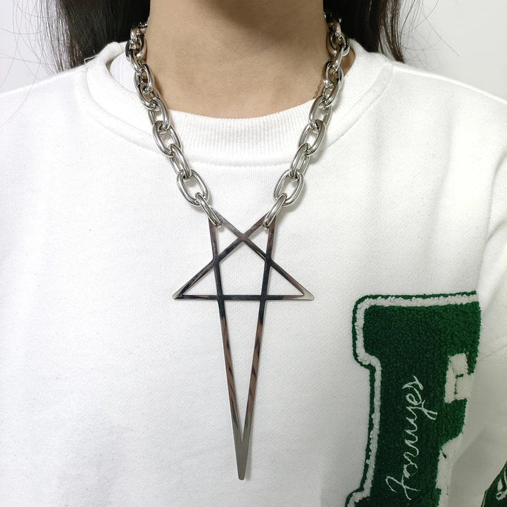 A person wearing a chunky stainless steel necklace with a large geometric star-shaped pendant, over a white sweatshirt with a green letter patch on the left side—a bold piece of streetwear jewelry.