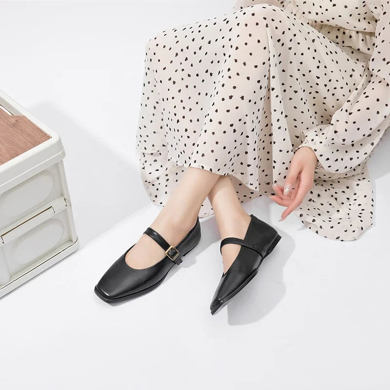 A person wearing a cream-colored dress with black heart patterns sits on the floor, showing off Augusta Leather Ballet Flats—stylish, sustainable shoes featuring a classic strap and buckle.