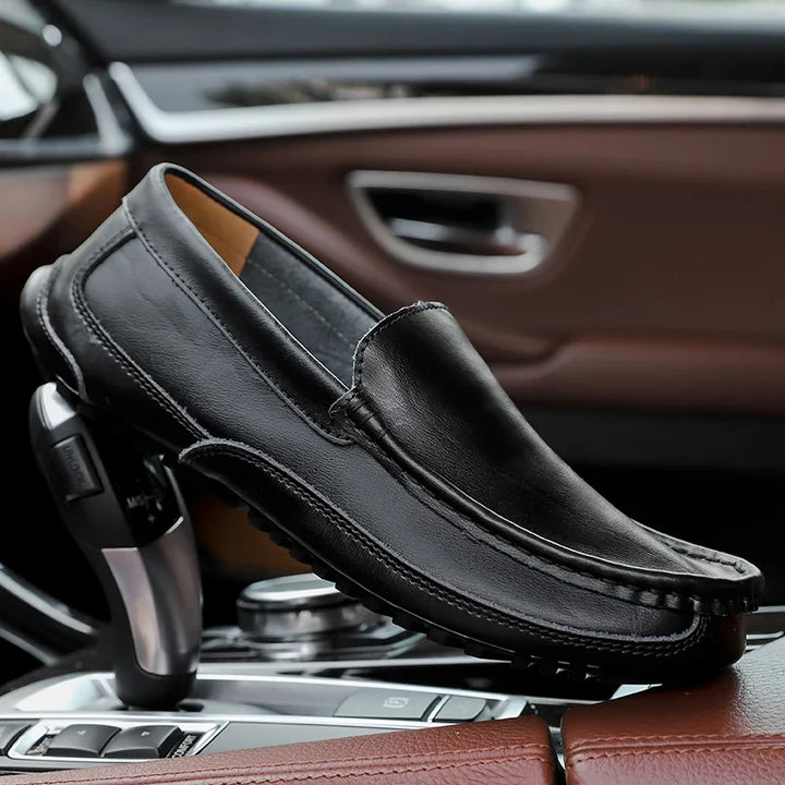A hand-stitched moccasin in black leather rests on the gear shift and console of a modern car with brown leather interior.
