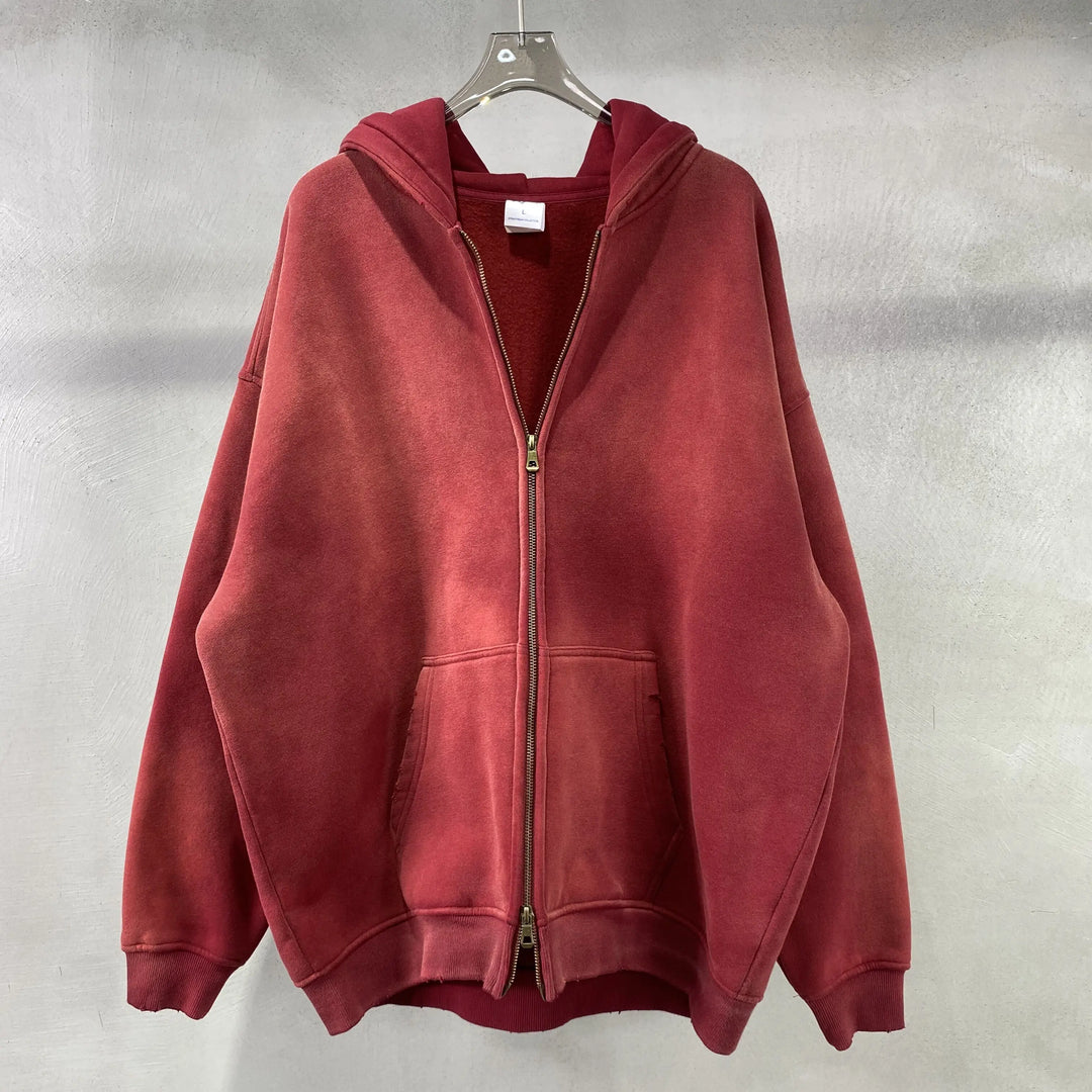 A spray-dyed vintage red zip-up hoodie with a relaxed fit and two front pockets hangs on a clear hanger against a light gray textured wall.