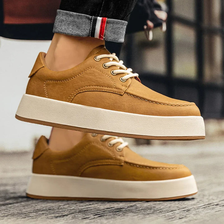 A person wearing light brown Men's Leather Casual Shoes with thick white soles stands on a wooden surface. The casual Italian designer shoes pair well with slightly rolled-up jeans, revealing a striped detail on the cuff.