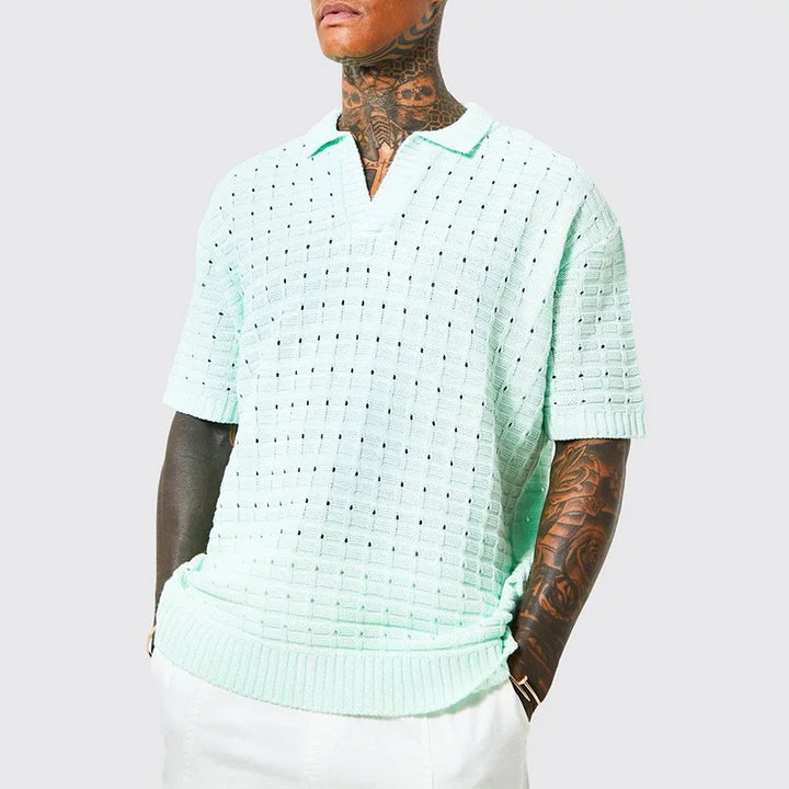 A person with tattoos on their neck and arms wears a light mint green, short-sleeved, summer shirt with a breathable knit design and textured, open-weave pattern, paired with white pants, standing against a plain light gray background.