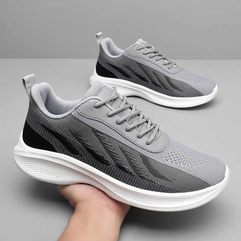 A pair of gray athletic sneakers with white soles and black accents. Featuring breathable mesh, one shoe is held up by a hand while the other rests on a smooth, gray surface against a plain background.