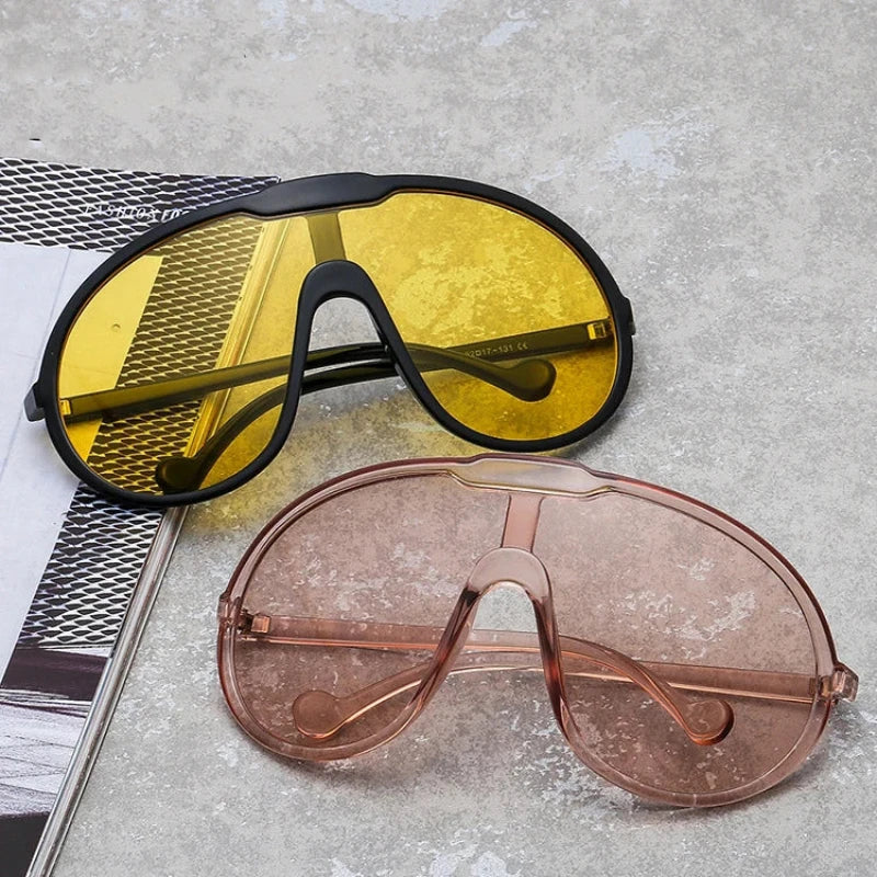 Two oversized sunglasses with UV400 protection lie on a gray surface next to an open magazine—one with yellow-tinted lenses and a black frame, the other featuring light pink-tinted lenses and a matching pink frame. Perfect for outdoor activities.
