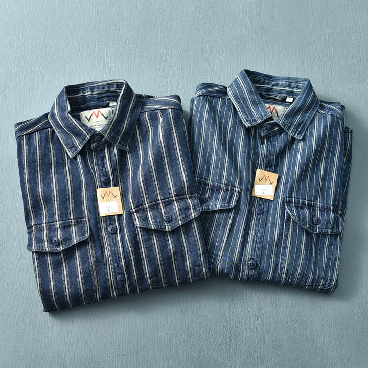 Two folded striped denim shirts with buttoned pockets, crafted from heavyweight cotton, are placed side by side on a light blue surface. Each shirt has a small tag attached near the collar.
