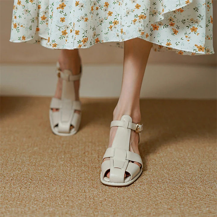 A person wearing handcrafted, cream-colored closed-toe sandals with straps and a buckle stands on a light brown carpet, paired with a white skirt featuring a yellow and orange floral pattern.