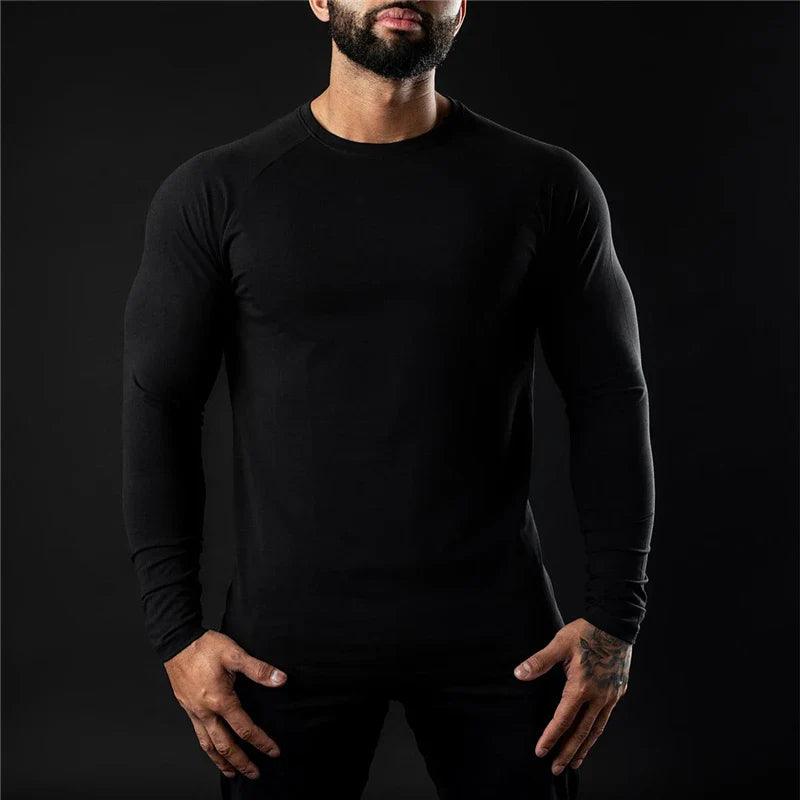 A man wearing a fitted moisture-wicking long sleeve shirt stands against a dark background, with his hands relaxed by his sides. His face is partially out of frame, showing only his beard and mouth.