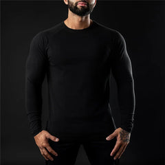 A man wearing a fitted moisture-wicking long sleeve shirt stands against a dark background, with his hands relaxed by his sides. His face is partially out of frame, showing only his beard and mouth.