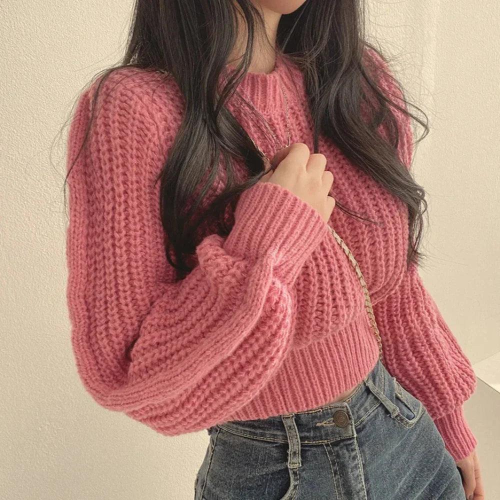 A person with long dark hair wearing a chunky pink knit sweater in a soft knit material and high-waisted blue jeans, holding a gold chain strap over their shoulder, stands against a light-colored wall.