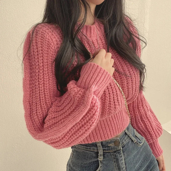 A person with long dark hair wearing a chunky pink knit sweater in a soft knit material and high-waisted blue jeans, holding a gold chain strap over their shoulder, stands against a light-colored wall.