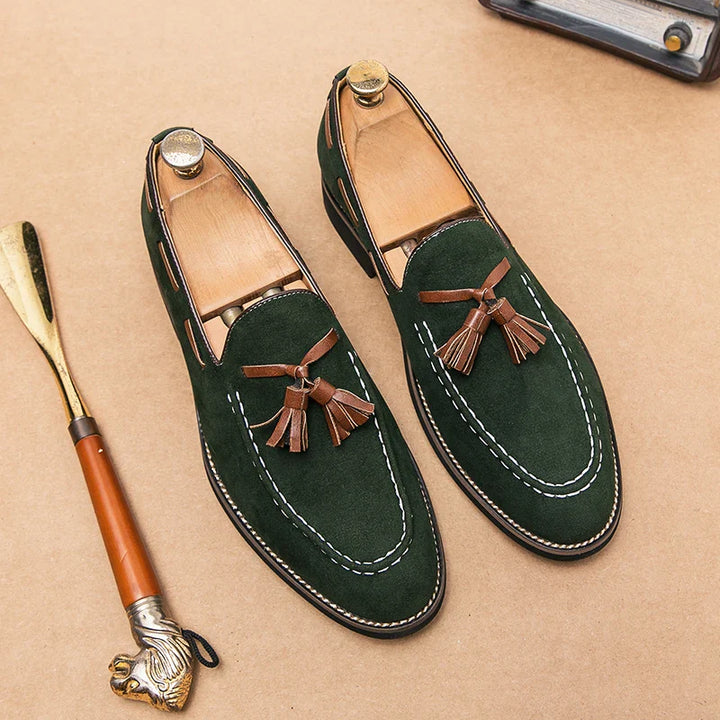 A pair of green suede tassel loafers with brown tassels and white stitching, displayed on a tan surface. Wooden shoe trees are inside each shoe, highlighting their durable construction. A gold-handled shoehorn lies nearby.