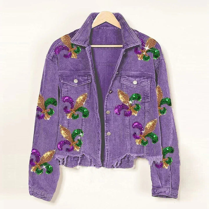 A purple corduroy jacket with a cropped relaxed fit hangs on a wooden hanger, adorned with green, gold, and purple sequin floral patches in fleur-de-lis patterns. The frayed hem and buttoned chest pockets add unique detail.