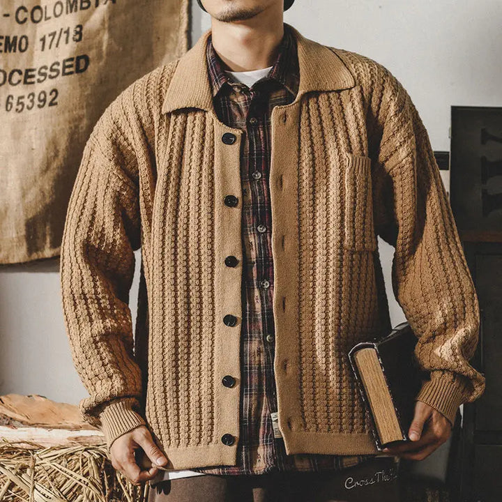 A man wearing a chunky knit beige cardigan over a plaid shirt holds a closed book in his left hand. The background features a burlap sack with printed text, highlighting the rustic charm and heritage aesthetic of the scene.