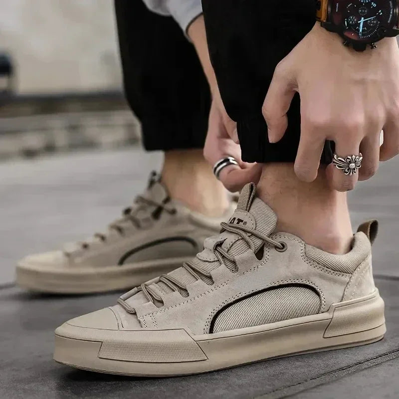A person wearing beige suede canvas shoes with a durable rubber outsole adjusts the cuff of their black pants. Rings and a watch accessorize their look, while the blurred background keeps the focus on their stylish footwear.