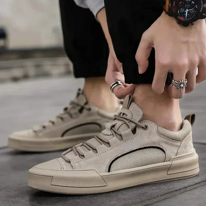 A person wearing beige suede canvas shoes with a durable rubber outsole adjusts the cuff of their black pants. Rings and a watch accessorize their look, while the blurred background keeps the focus on their stylish footwear.