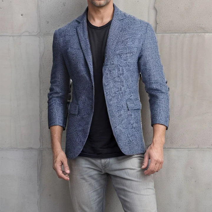 A man stands against a gray concrete wall wearing a textured blue blazer over a black t-shirt and light gray tapered fit jeans. His head is not visible in the image.