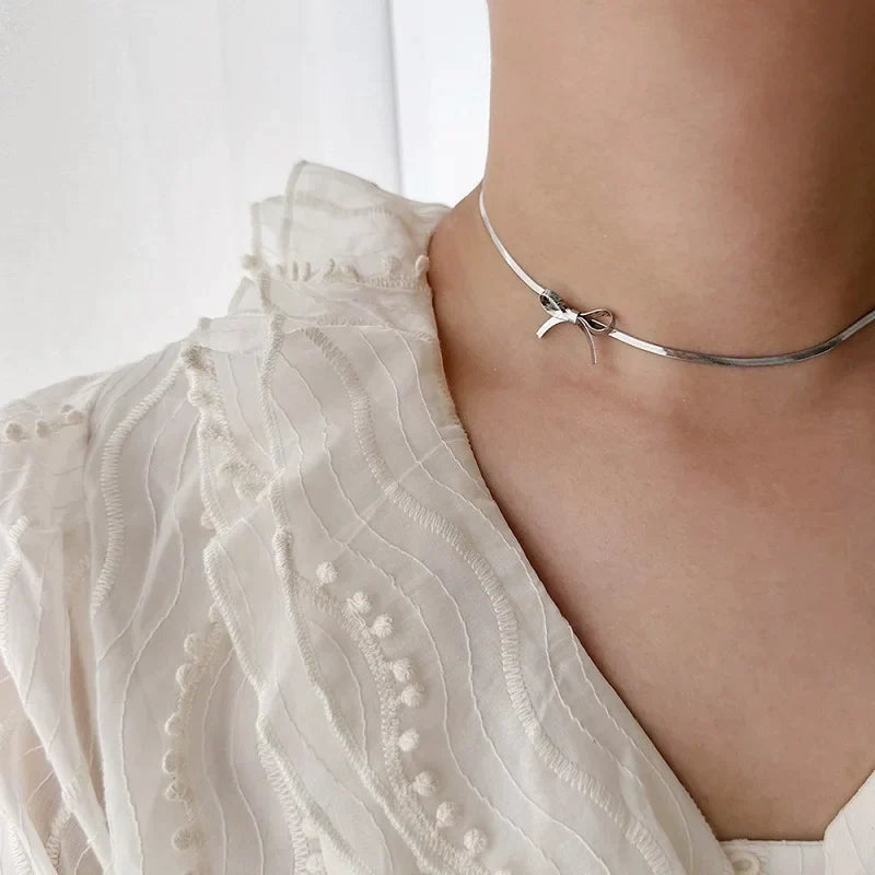 A person wearing a textured cream-colored blouse with ruffles and embroidered details, paired with a minimalist jewelry piece: a thin silver bow necklace that adds an elegant touch.