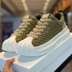 A hand holds a white shoebox with olive-green woven upper sneakers featuring thick beige laces, a chunky cushioned sole, and a rounded toe, displayed indoors under bright lighting.