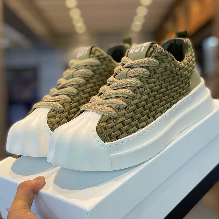 A hand holds a white shoebox with olive-green woven upper sneakers featuring thick beige laces, a chunky cushioned sole, and a rounded toe, displayed indoors under bright lighting.