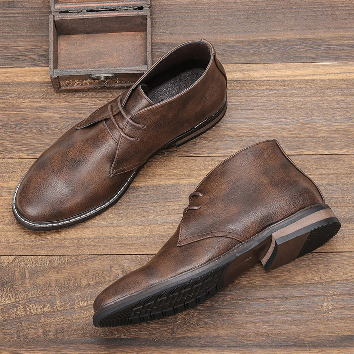 A pair of brown leather After-Ride Chukka dress shoes displayed on a wooden surface, with one shoe upright and the other lying on its side to highlight the sole and side details.