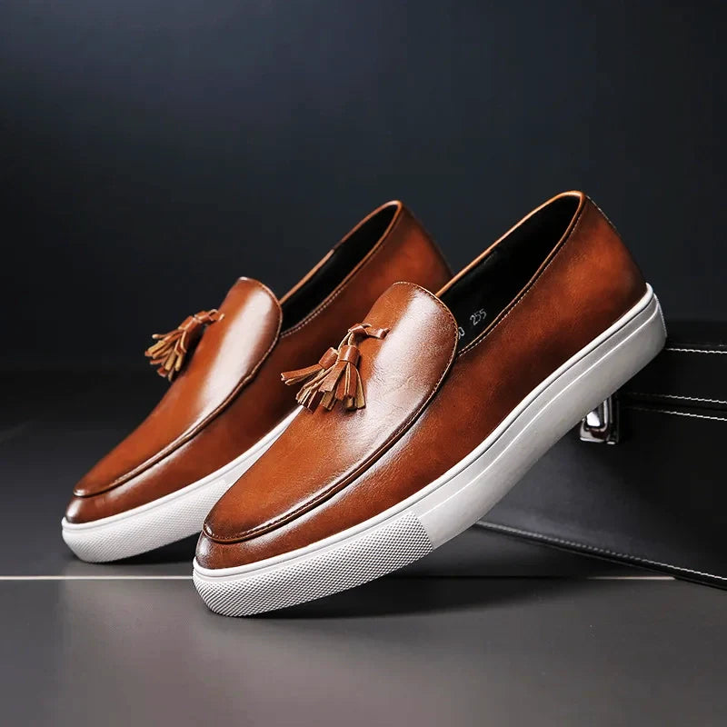 A pair of luxury leather loafers with tassels and white rubber soles, featuring a cushioned insole for comfort, displayed against a dark background with one shoe propped up on a black box.