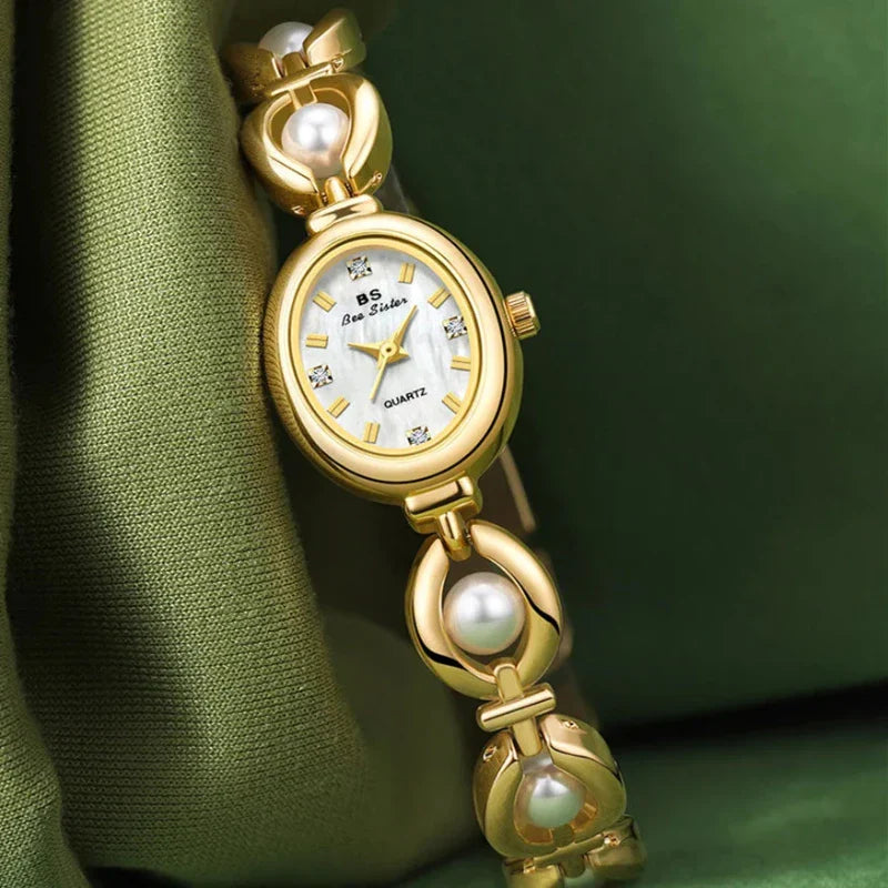 A gold womens wristwatch with a white oval face, gemstone hour markers, and a bracelet-style band adorned with pearl accents, featuring precision quartz movement—elegantly displayed on green fabric.