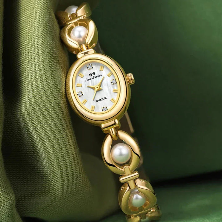 A gold womens wristwatch with a white oval face, gemstone hour markers, and a bracelet-style band adorned with pearl accents, featuring precision quartz movement—elegantly displayed on green fabric.