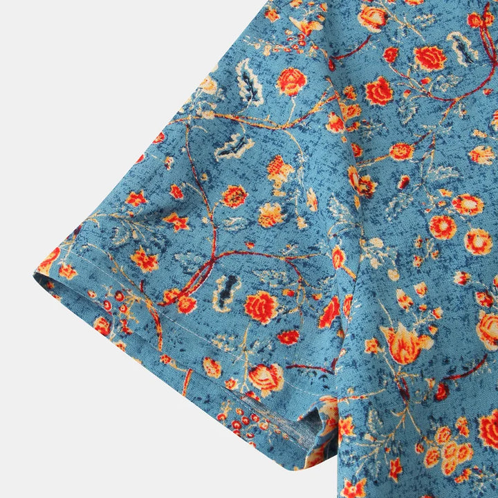 Close-up of a blue cotton shirt with a colorful floral pattern in red, orange, and yellow, showing the sleeve. The flowers and vines are scattered over the textured background, adding charm to this classic design.