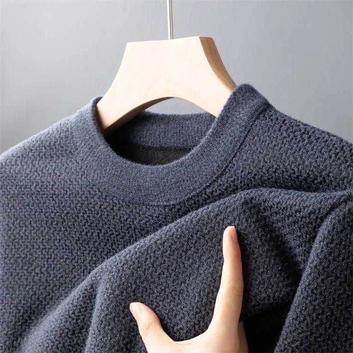 A hand touches a relaxed fit sweater, crafted from high-quality cotton, its dark gray knit hanging on a wooden hanger against a plain gray background.