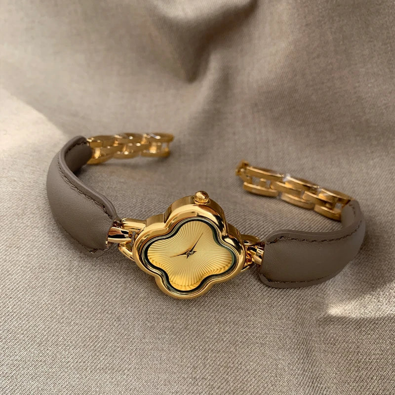 A luxury gold-plated clover dial watch with a brown genuine leather strap and gold link band, displayed on a beige fabric background. The simple, elegant face features a unique clover shape without numbers.