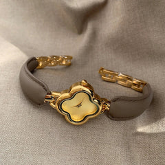A luxury gold-plated clover dial watch with a brown genuine leather strap and gold link band, displayed on a beige fabric background. The simple, elegant face features a unique clover shape without numbers.