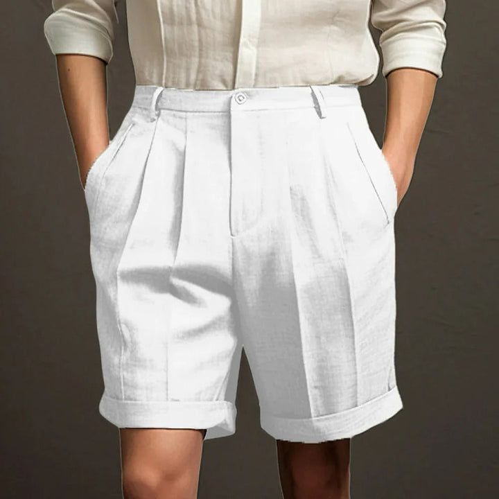 A person wearing a white button-up shirt with the sleeves rolled up and high-waisted, breathable linen shorts with front pleats and cuffed hems, standing with hands in their pockets—perfect for a relaxed summer look.