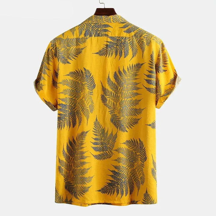 A classic design cotton shirt with short sleeves in yellow, featuring large dark green and blue fern leaves, is shown from the back on a hanger against a plain white background—perfect for a casual brunch.