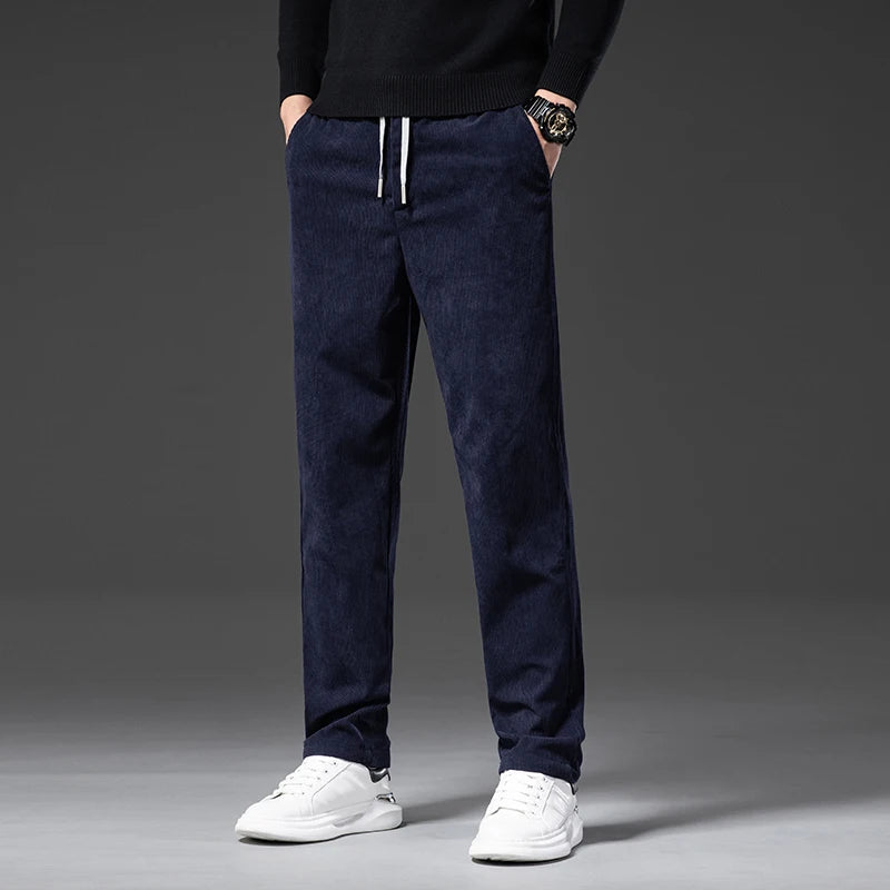 A person wearing navy blue men's smart casual pants with a drawstring, a black sweater, white sneakers, and a watch stands against a plain dark background. The photo is cropped at the shoulders, showing only the lower body.