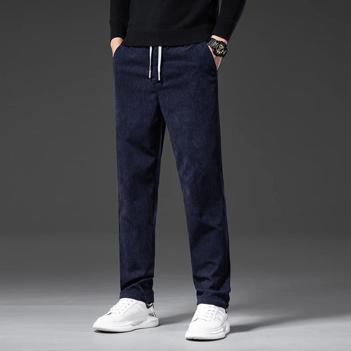 A person wearing navy blue men's smart casual pants with a drawstring, a black sweater, white sneakers, and a watch stands against a plain dark background. The photo is cropped at the shoulders, showing only the lower body.