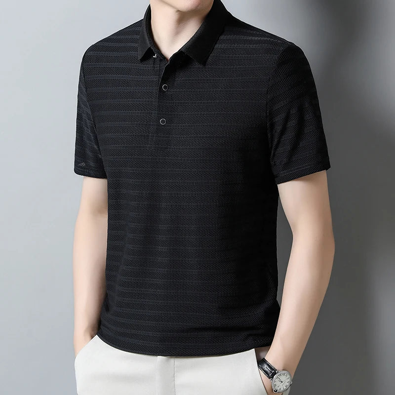 A person wearing a short-sleeved, black, textured classic polo with beige pants and a wristwatch, standing against a plain gray background.