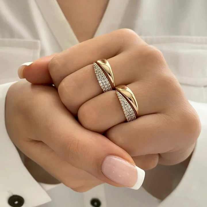 A close-up of hands wearing two gold rings, one with a twisted design and pavé diamond accents, set against a white shirt. The nails are manicured with pale pink polish and white tips.