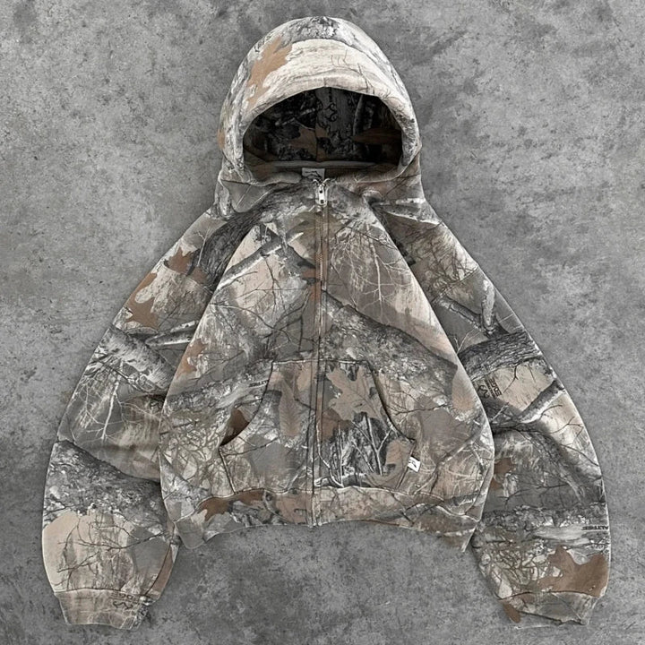 A hooded zip-up jacket with a gray and brown camo pattern is laid flat on a textured gray surface. Featuring long sleeves, ribbed cuffs, and an oversized fit, this jacket is crafted from a soft cotton blend for all-day comfort.