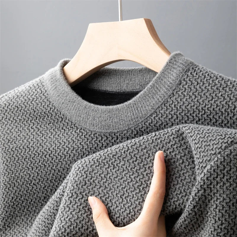 A hand is touching the sleeve of a relaxed fit sweater, crafted in textured, light gray knit, hanging on a wooden hanger against a plain gray background.