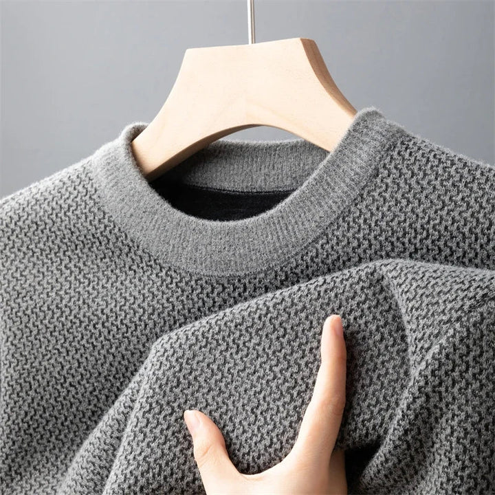 A hand is touching the sleeve of a relaxed fit sweater, crafted in textured, light gray knit, hanging on a wooden hanger against a plain gray background.