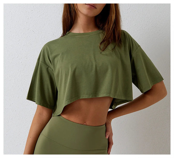 A woman in a relaxed fit, olive green cotton crop top and matching high-rise pants stands against a light-colored wall. Her face is partially out of the frame, and one hand rests on her hip.