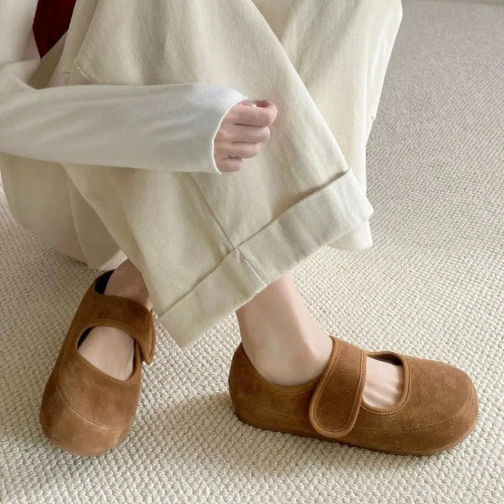 Person wearing cream-colored pants and a long-sleeved top crouches on a textured light carpet, showcasing tan suede Mary Janes—stylish women casual shoes with a Velcro strap, perfect for everyday comfort.