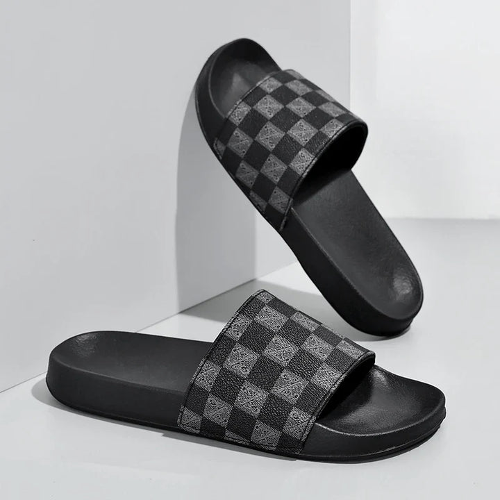 A pair of black streetwear slides with a gray and black checkerboard pattern on the straps, displayed against a white background. One checkered sandal leans against the wall while the other rests flat on the surface.