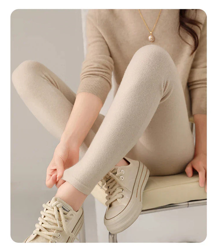 A person dressed in a beige sweater and high-waisted leggings sits on a chair, crossing one leg. They wear beige sneakers, and a gold necklace with a pearl pendant is visible. Their face is not shown.
