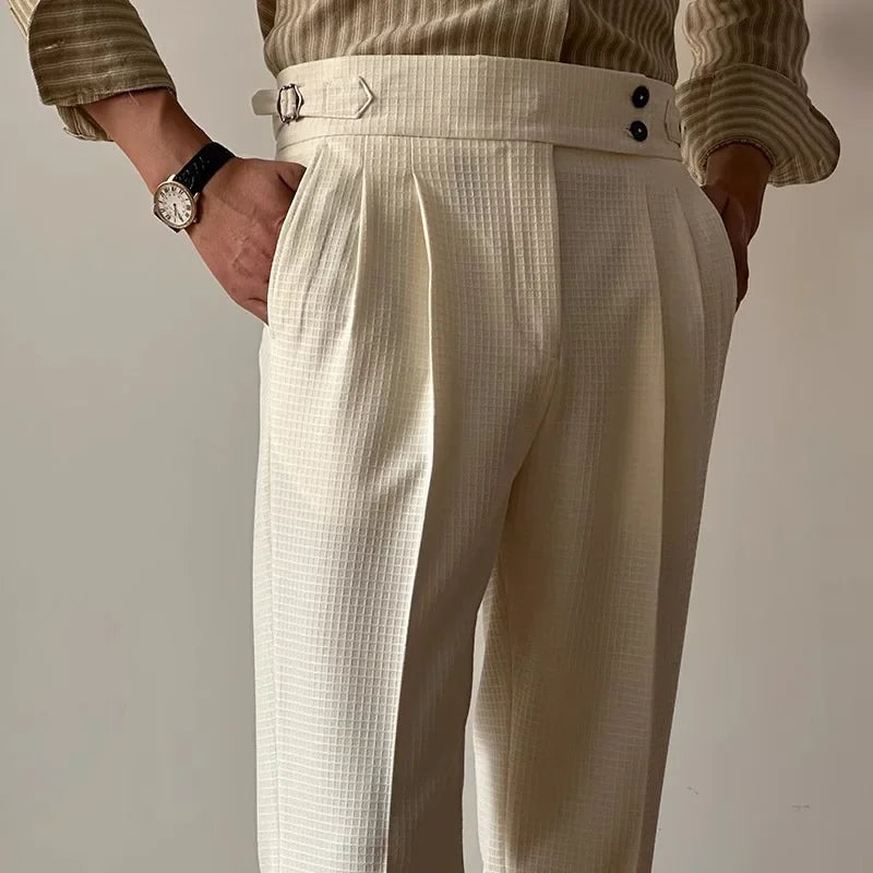 A person stands with one hand in their pocket, wearing cream-colored Havana Fit Slacks with side buttons and a beige striped shirt. The high-waisted pleated trousers are paired with a watch on their left wrist.