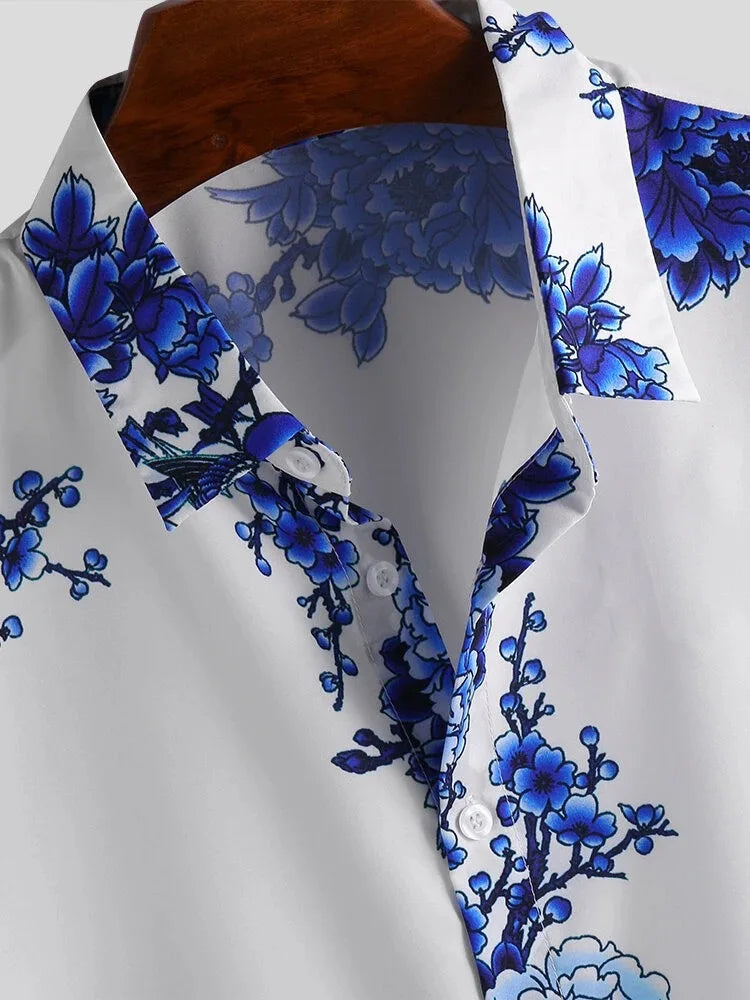 A white collared floral design shirt featuring a bold blue print along the collar, shoulders, and chest, displayed on a wooden hanger. Partially unbuttoned and crafted in a cotton-silk blend, its perfect for a summer-ready look.