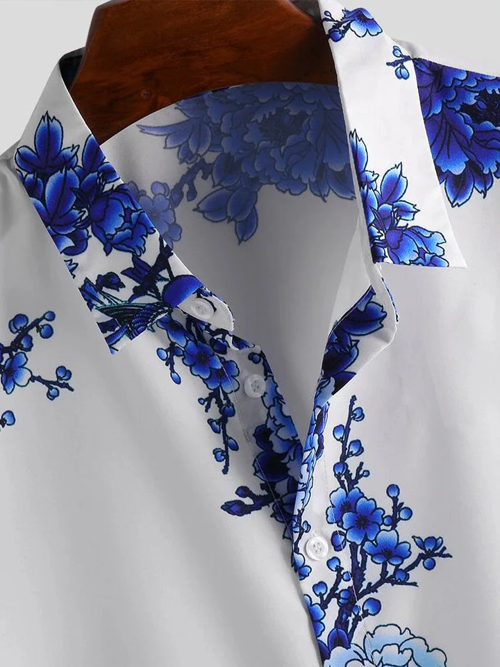 A white collared floral design shirt featuring a bold blue print along the collar, shoulders, and chest, displayed on a wooden hanger. Partially unbuttoned and crafted in a cotton-silk blend, its perfect for a summer-ready look.