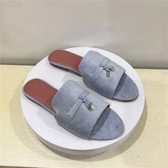 A pair of light gray suede slip-on sandals with a cushioned insole and brown insoles, displayed on a round white platform against a beige background. Each sandal features a decorative bow and small metallic charm on the upper.