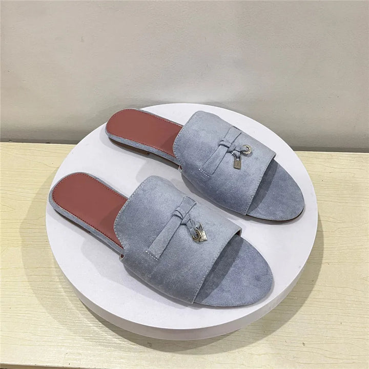 A pair of light gray suede slip-on sandals with a cushioned insole and brown insoles, displayed on a round white platform against a beige background. Each sandal features a decorative bow and small metallic charm on the upper.