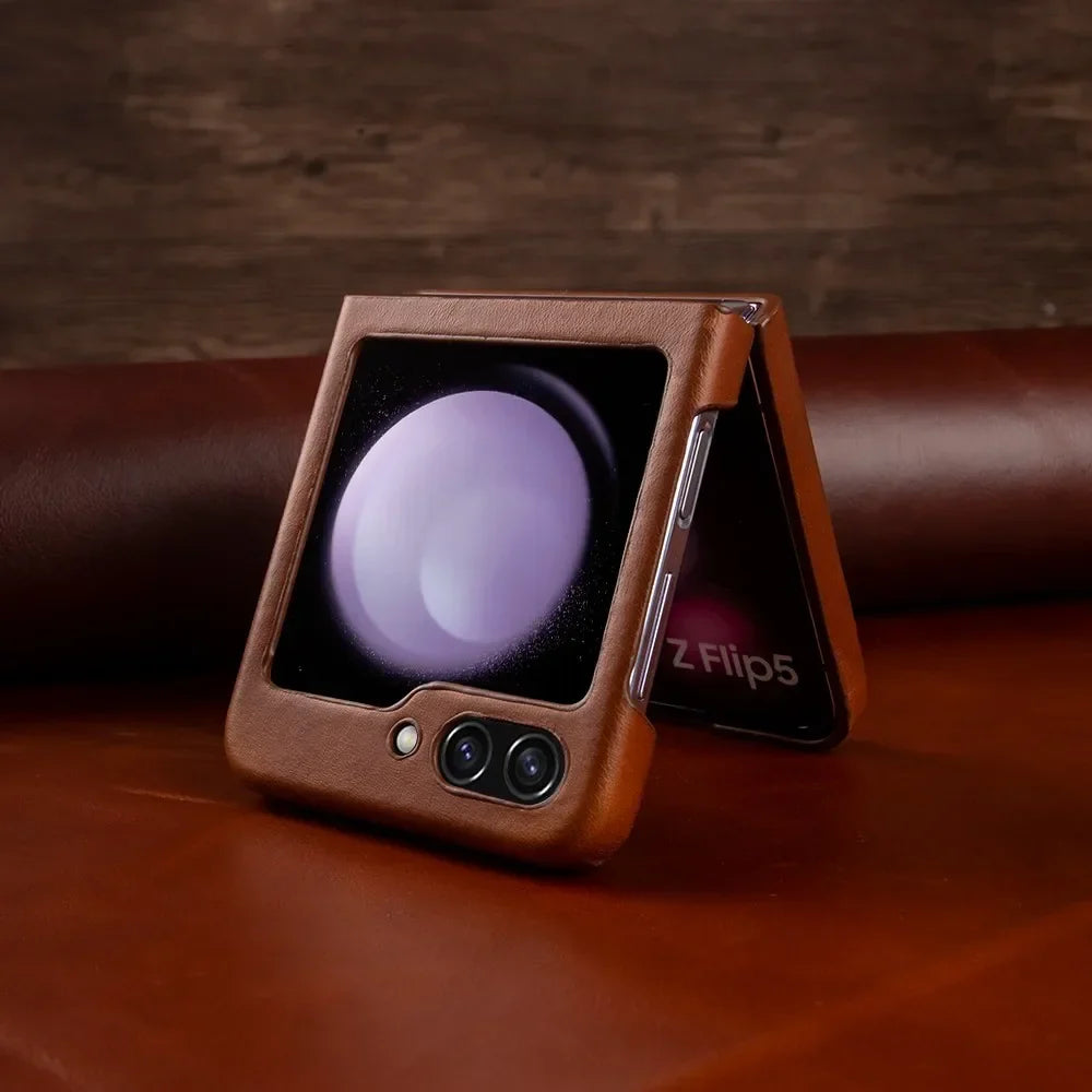 A Samsung Galaxy Z Flip5 smartphone in a genuine leather case is partially folded and resting on a brown surface. The outer display shows a circular graphic, and the dual rear cameras are visible for stylish shockproof protection.